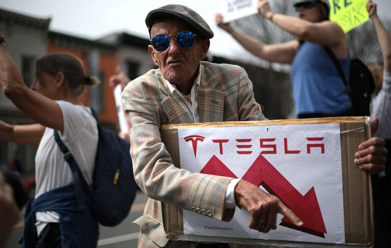 Tesla protester holds a sign showing Tesla's stock price going down.