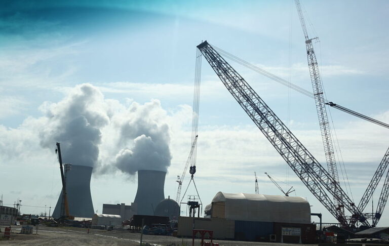Construction cranes in action in the foreground, while steam spills from two cooling towards in the background.
