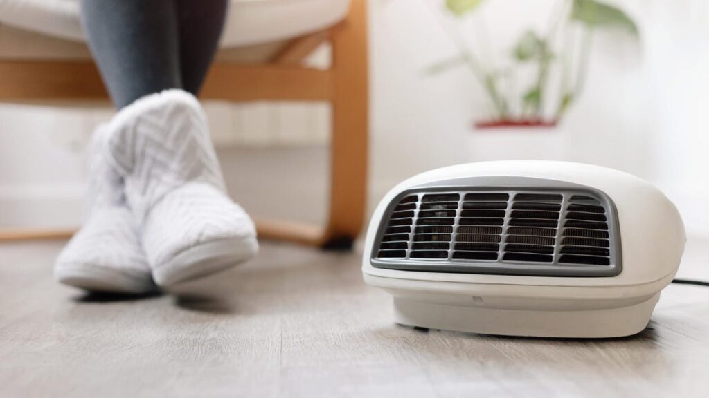 A person in warm boots sits in front of a space heater on the ground.