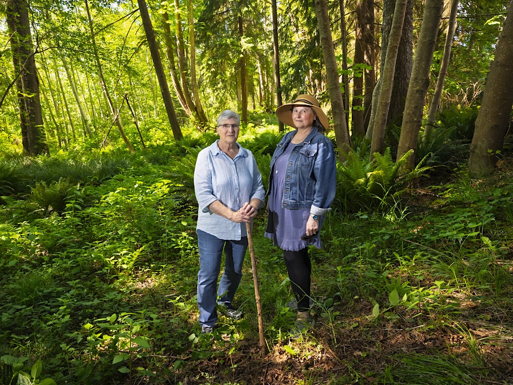 Two women stand in a lush urban forest, surrounded by ferns and trees.