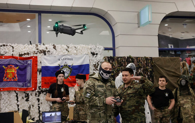 Shaman, a 19-year-old drone operator for the Russian military, flies a quadcopter drone during a demonstration event organized by members of the Berkut Military-Sports Cossack Club in a shopping centre in Voronezh, Russia on January 24, 2026. Shaman is wearing a face mask and military uniform with a camo pattern while standing next to another man in similar attire, controlling a quadcopter drone hovering in the foreground. Behind them are two girls and a boy wearing military camo-style pants and black t-shirts, along with the Russian flag.