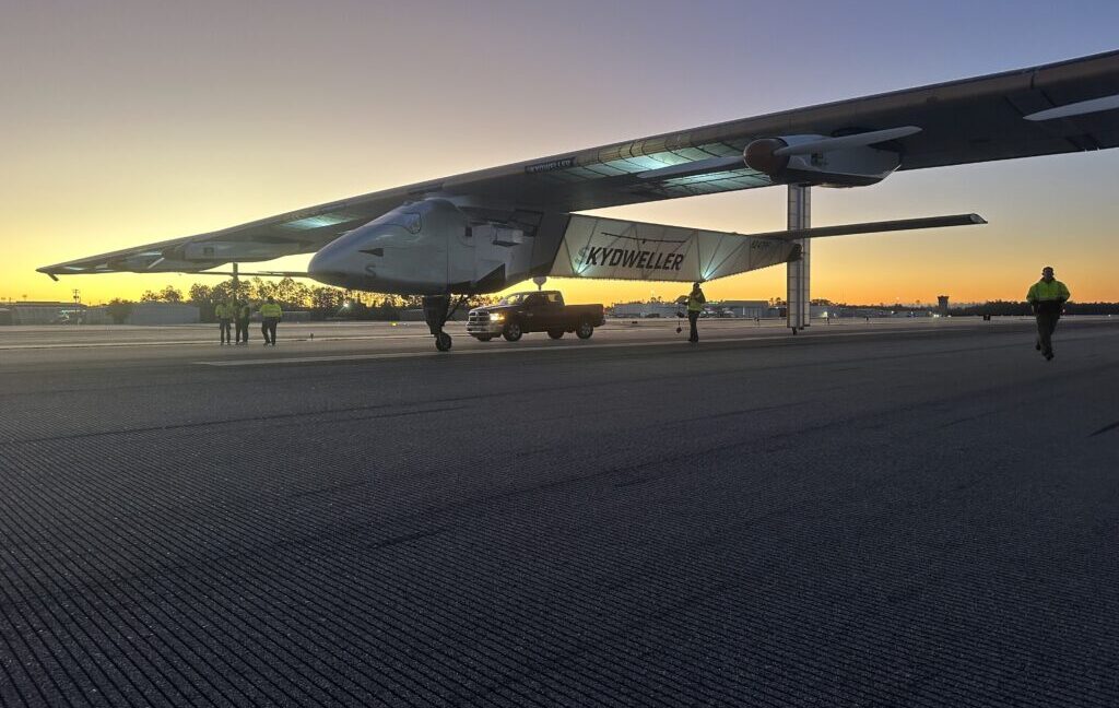 A predawn image of the Skydweller Aero drone with jumbo jet sized wings sitting on a runway or taxiway. Three people stand near the aircraft's right wing while another person is walking near the left wing. A pickup truck is parked alongside the aircraft.