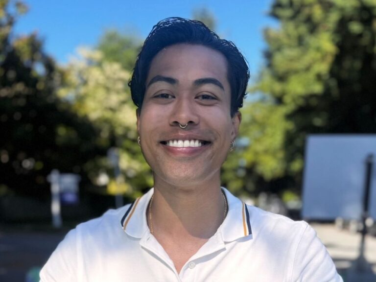 A man with medium-skin, wearing a white polo shirt, smiles for the camera on a sunny day in a place with some deciduous trees.