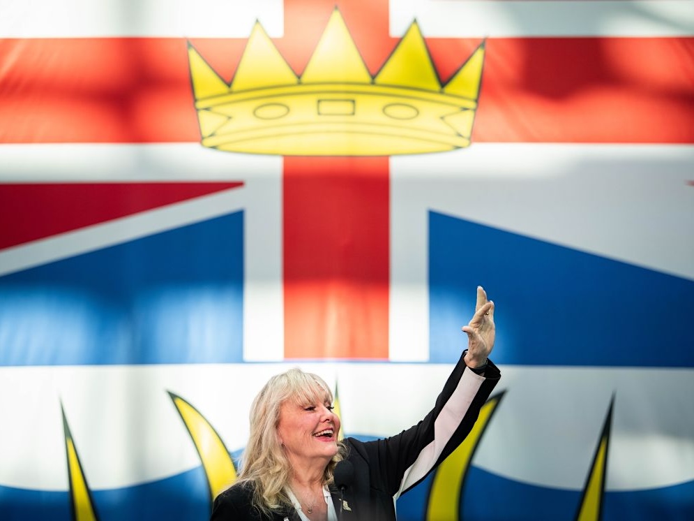 A woman in her 70s with shoulder-length blond hair smiles and waves. A large British Columbia flag can be seen behind her, with the crown on the flag positioned above her head.
