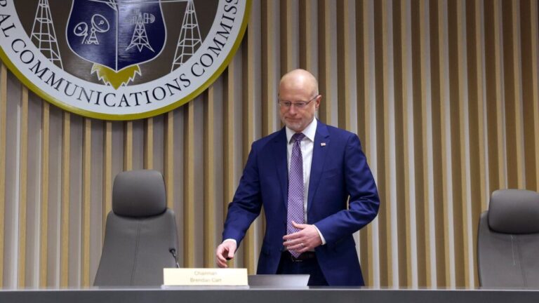 FCC Chairman Brendan Carr prepares to sit down for a meeting. A large FCC crest is seen on the wall above him.