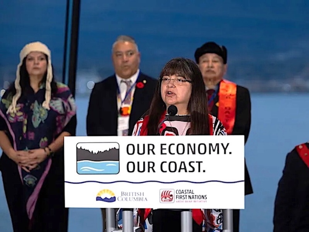 A middle-aged Indigenous woman stands at a podium that says &lsquo;Our Economy, Our Coast&rsquo; with other Indigenous people behind her.