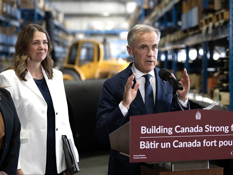 Mark Carney gestures with his hands while speaking at a lectern inside a warehouse. Danielle Smith stands on the left, looking on.