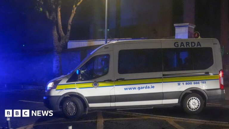 A white Garda van with a yellow stripe along its side pulls out onto a main road on a dark night.