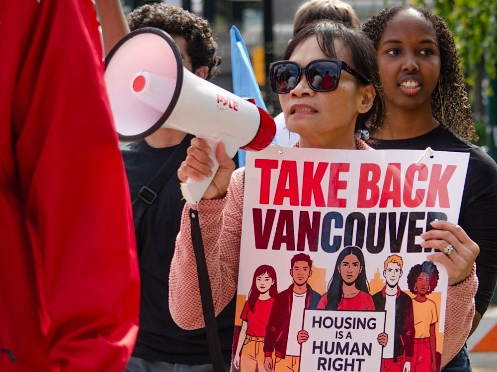 A woman wearing sunglasses holds a sign that says, &lsquo;Take Back Vancouver&rsquo; and &lsquo;Housing as a Human Right&rsquo; at a political rally on the grounds of Vancouver City Hall.