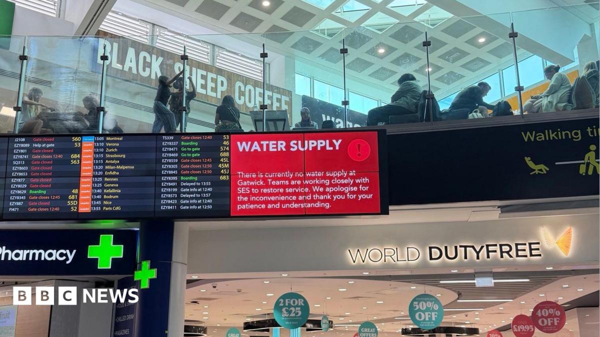 A red sign alerts passengers to the water supply issue on a screen in a terminal building. A World Duty Free sign is underneath and a pharmacy sign is nearby. A further screen shows flight information.