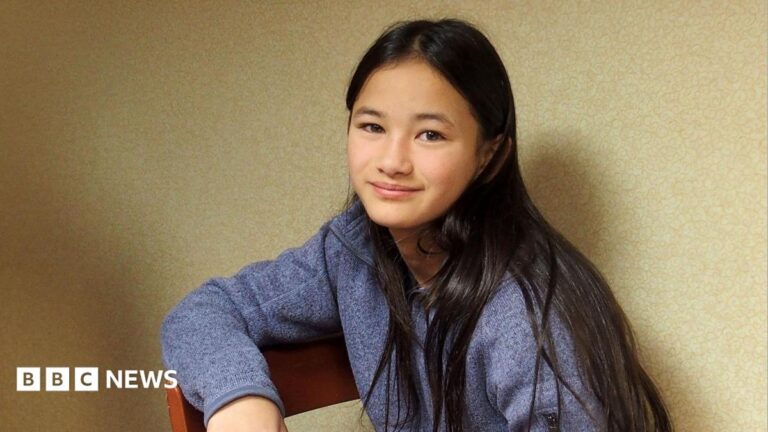 Kaylee Hottle sits on a wooden chair and looks into the camera. She wears a blue jumper and has long dark hair.