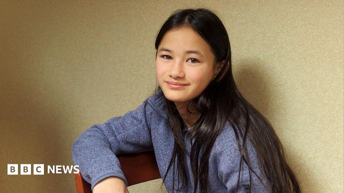 Kaylee Hottle sits on a wooden chair and looks into the camera. She wears a blue jumper and has long dark hair.