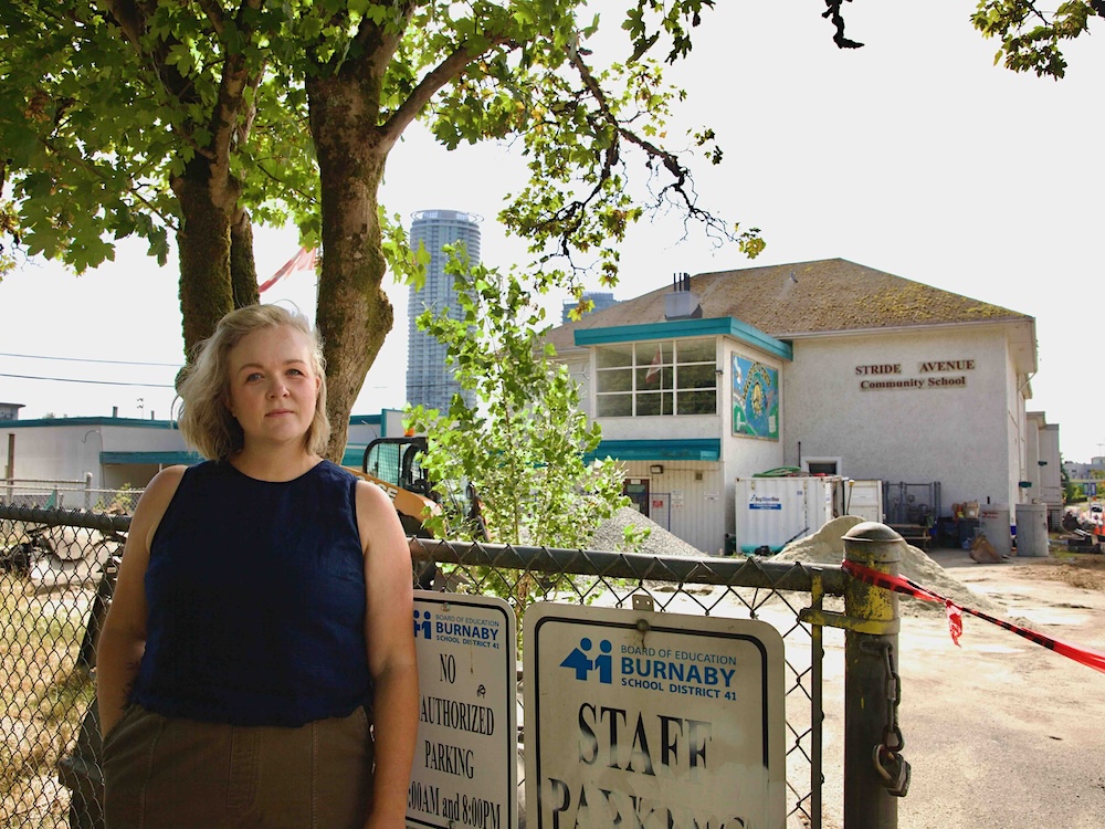 Jamie Owen is standing against a fence in front of a white two-storey school surrounded by construction. She has shoulder-length blond hair and a dark blue sleeveless top.