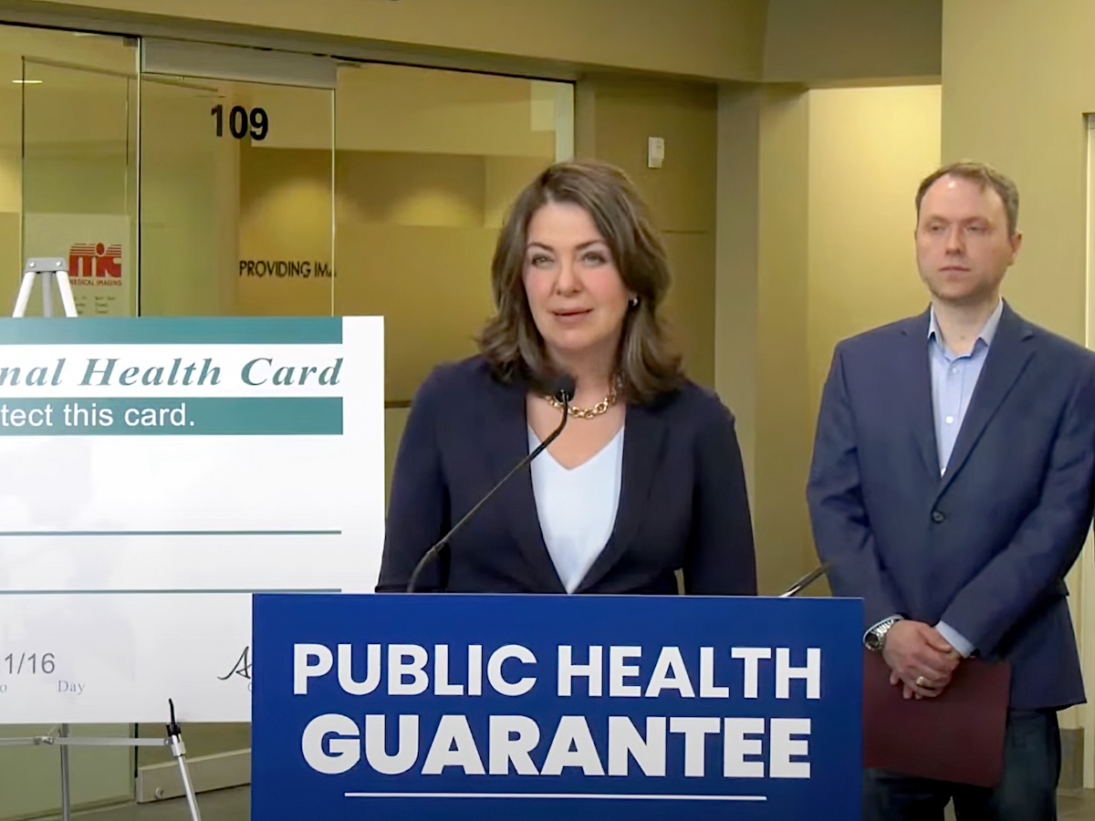 Alberta Premier Danielle Smith, a middle-aged white woman with shoulder-length brown hair, wearing a suit, stands behind a dais with a sign on it saying ‘Public Health Guarantee.’