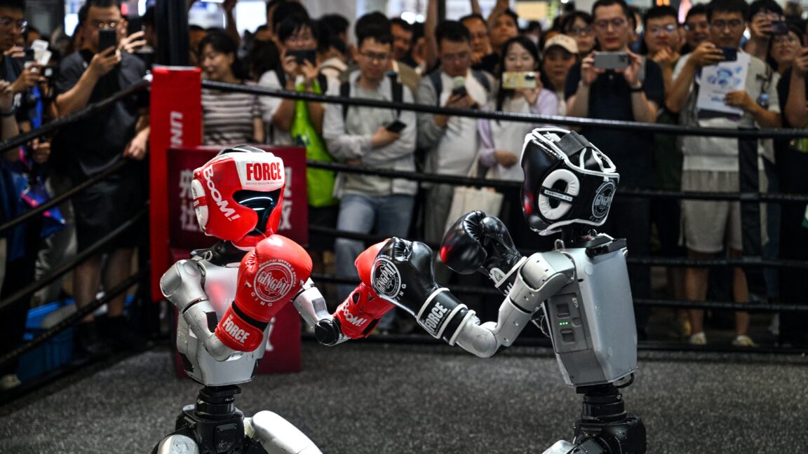Humanoid robots controlled by a remote control by Unitree Robotics take part in a fight during the World Artificial Intelligence Conference (WAIC) in Shanghai on July 18, 2026. The foreground shows one robot on the left wearing a red helmet and boxing gloves and one robot on the right wearing black gear. The background shows a crowd of onlookers standing behind a boxing ring's ropes.