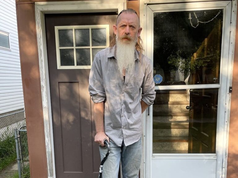 A man with light skin tone and a long grey beard stands on his porch, using a cane.