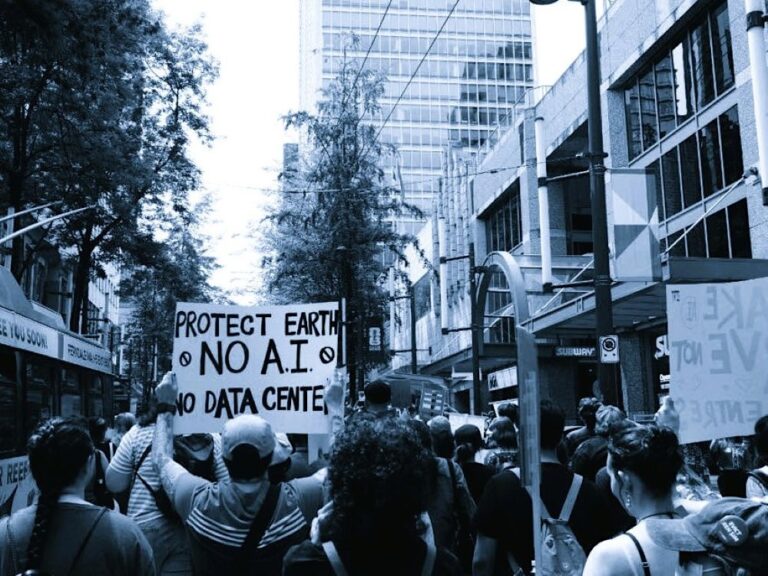 A black and white photo of a group of protesters from behind. One holds up a sign that says, ‘Protect Earth. No AI. No Data Centre.’