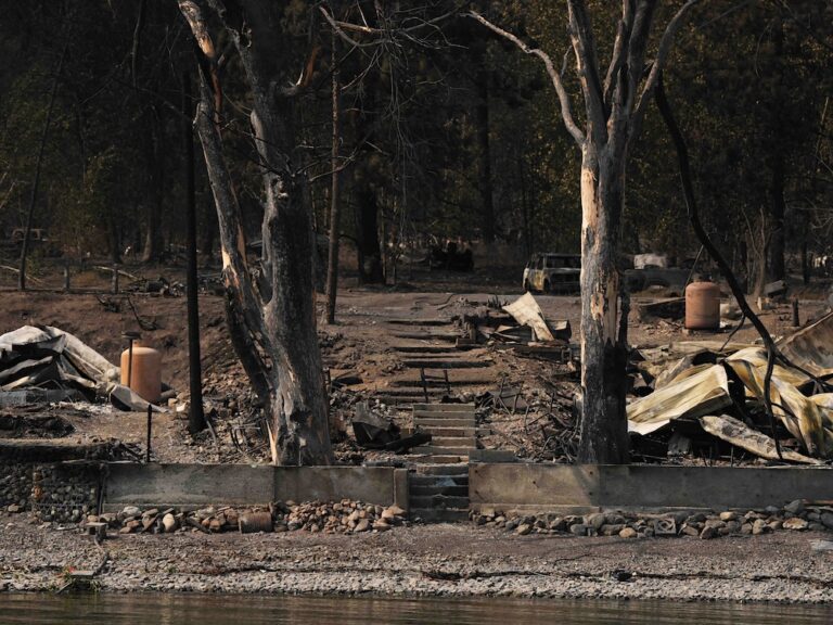 Burned trees and rubble from two burned structures can be seen on the shore of a lake.