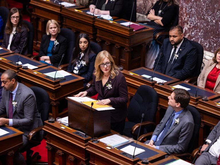A middle-aged woman with light skin and long blond hair wearing a suit stands at her desk in B.C.’s legislature chamber. Other MLAs wearing suits and other formal business attire sit around her.