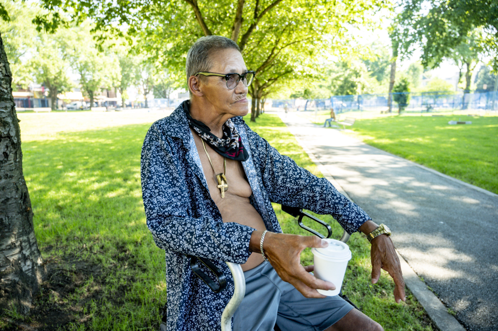 A man with short greying hair and medium-light skin tone sits on a rollator in a park. He is wearing a bandana and an unbuttoned shirt and holding a coffee.