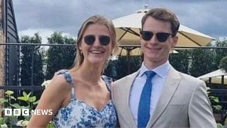 Alexander Cromie and Marie Ebert stand arm in arm and smile. Alexander is wearing a tan suit and sunglasses. Marie is also wearing sunglasses and a floral dress.