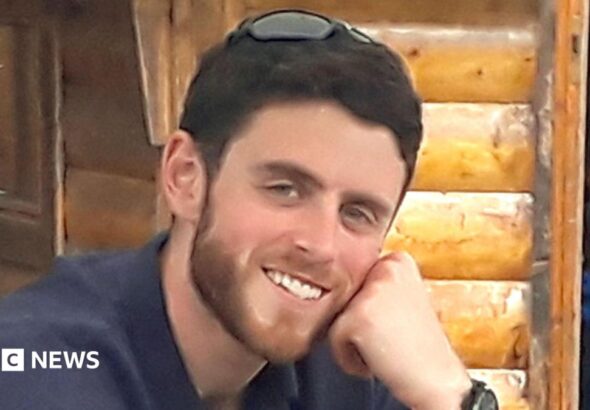 a man with short brown hair and a slight beard wearing a shirt and smiling at the camera.