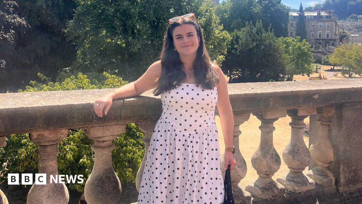 Ella Kipling, a young woman with long dark hair, smiles at the camera. She is wearing a long, white dress with black polkadots and is standing in front of a large park with old buildings in the background.