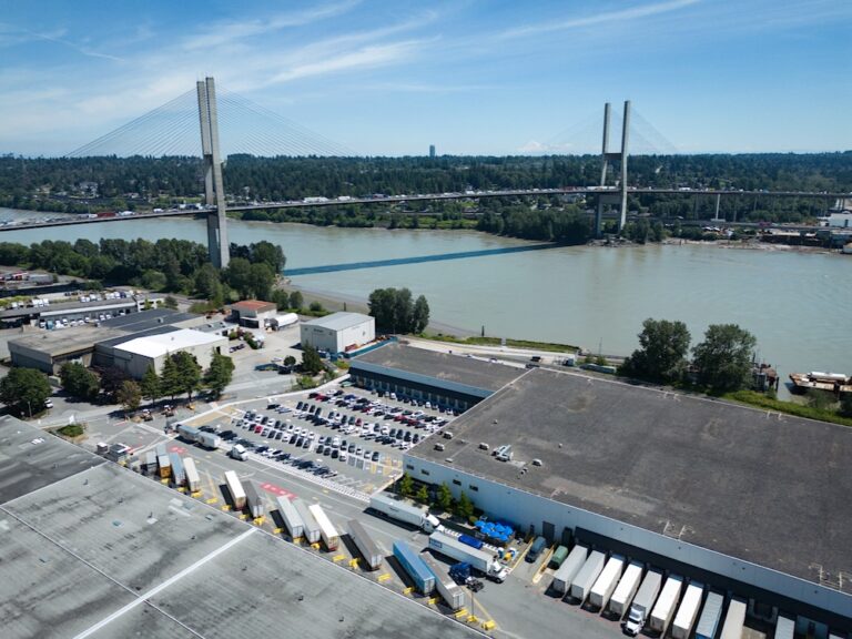 An aerial view of a large warehouse building surrounded by a parking lot, with a river and bridge in the background.