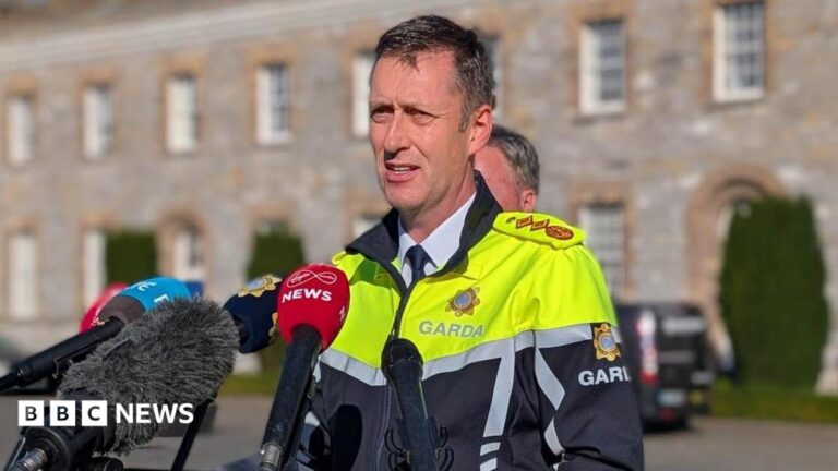 Garda Commissioner Justin Kelly syanding in front of microphones. He has short dark hair, wearing a white shirt, navy tie and high viz garda jacket.