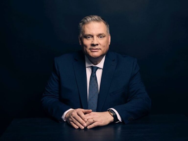 A middle-aged man with light skin and short ash-blond hair, wearing a dark suit and tie, sits with his hands in front of him.