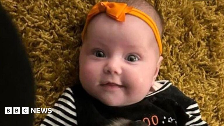 Dahlia-Rose Gartshore, a three-month-old baby girl looking at the camera, with a slight smile. She has an orange band on her head with a bow.
