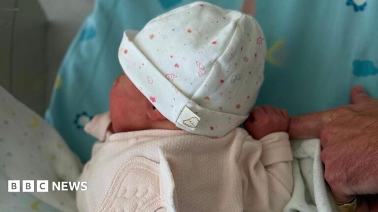 A baby wearing a pink sleepsuit and a white hat with pink decoration. The finger of an older adult's hand is in the picture with the baby clasping it