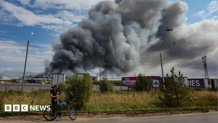 Large fire engulfs a market area at night, sending towering flames and thick smoke into the sky above nearby buildings, streets, and overhead power lines.