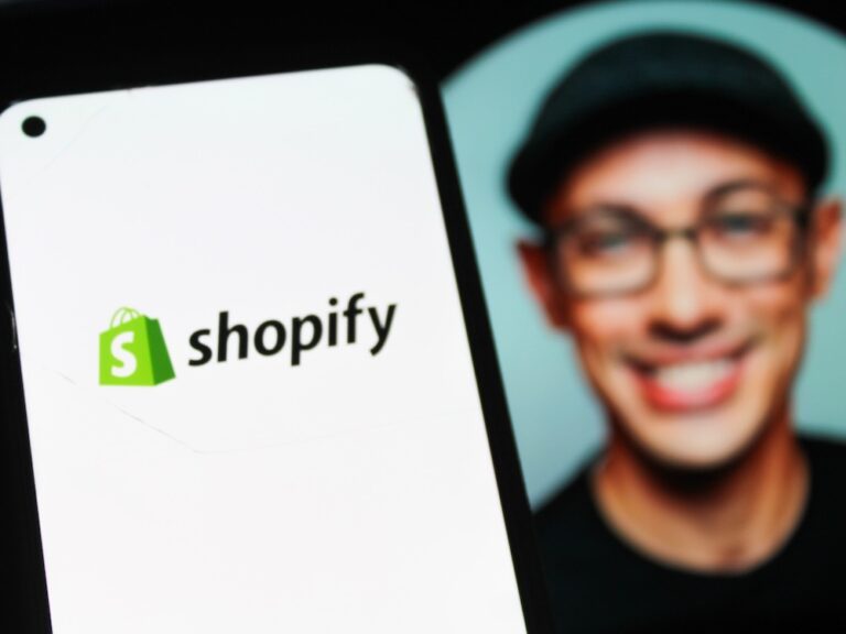 The Shopify logo displayed on a smartphone screen. Shopify CEO Tobi Lütke’s Instagram photo can be seen in the background out of focus: a middle-aged man wearing glasses and a cloth cap and smiling broadly.