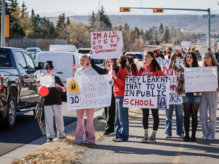 A group of striking teachers stand on a sidewalk holding protest signs.