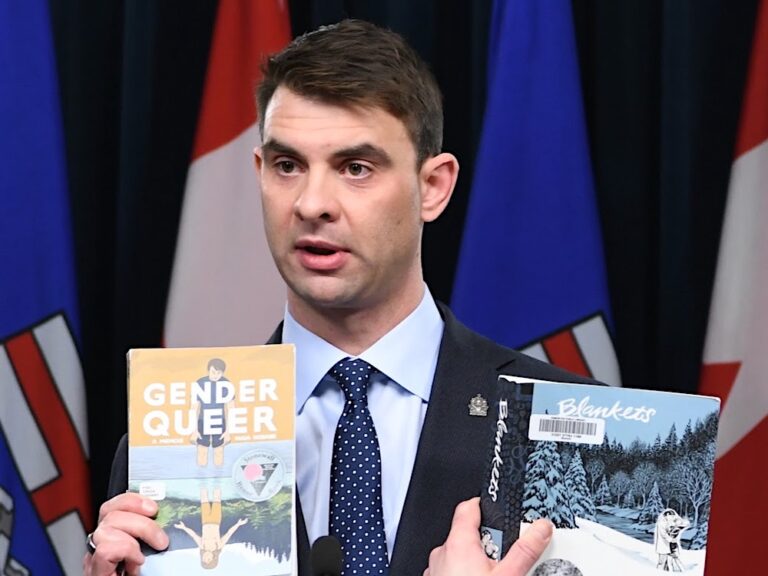 A 40-ish man with light-toned skin and dark hair is wearing a suit and holding two books titled ‘Gender Queer’ and ‘Blankets.’