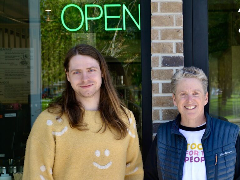 Chance McClendon, left, wears a yellow shirt with smiley faces on it. He stands beside Jenny Farkas, who is wearing a blue vest and a shirt with the words ‘Tools for the People’ on it. They are in front of a door and brick wall with an ‘Open’ sign.