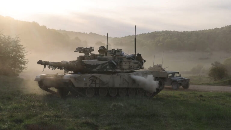 A US Army tank can be seen in profile with its main gun facing to the left. The background shows another US Army vehicle driving by with dust in the air. The scene took place during Exercise Combined Resolve held in Germany from April 9 to May 10, 2026.