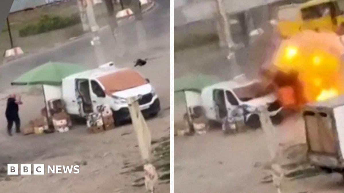A screengrab of drone footage showing crosshair sights and an overhead view of white car on a street at the moment when an explosive detonates on its roof