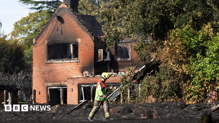 A firefighter wearing a helmet and hi-vis walks in front of a burnt out red-brick house.