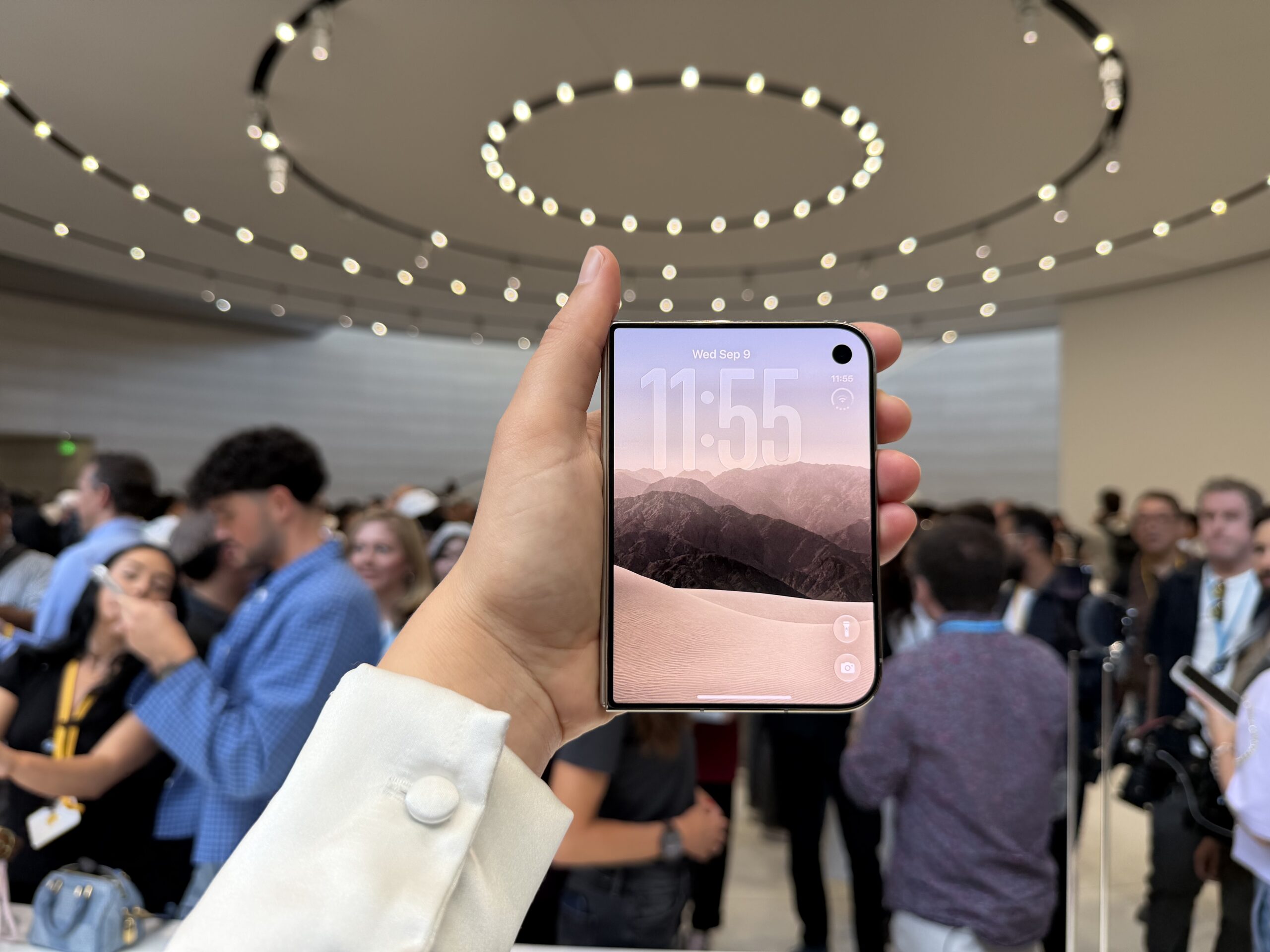 hand holding the iPhone Duo in a busy room, with the front screen showing mountains and the time 11:55