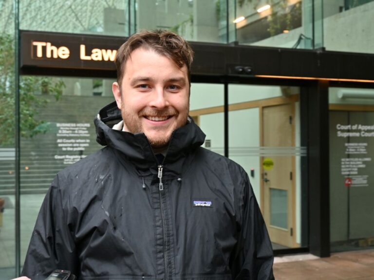A light skinned man wearing a dark-coloured rain jacket smiles at the camera.