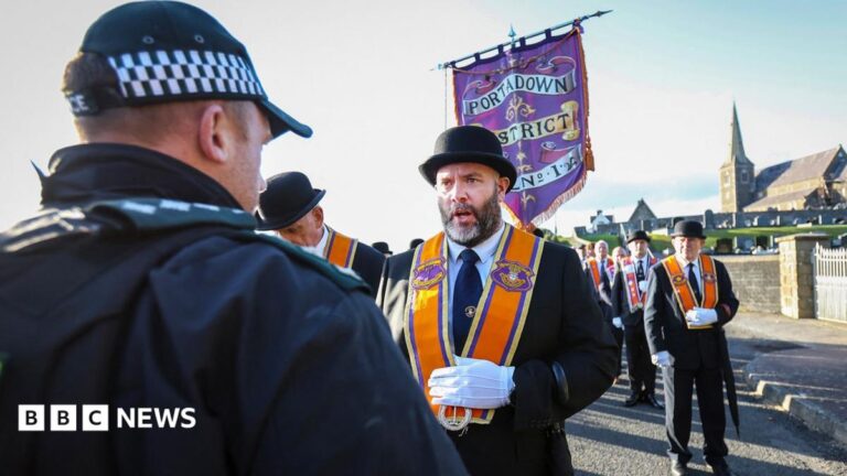 An Orange Order member talking to a police officer wearing a black suit, black hat and an orange sash. The rest of the lodge members are behind him with the church in the background. The police man is wearing a black and white checked hat with a black coat with green PSNI lapels.