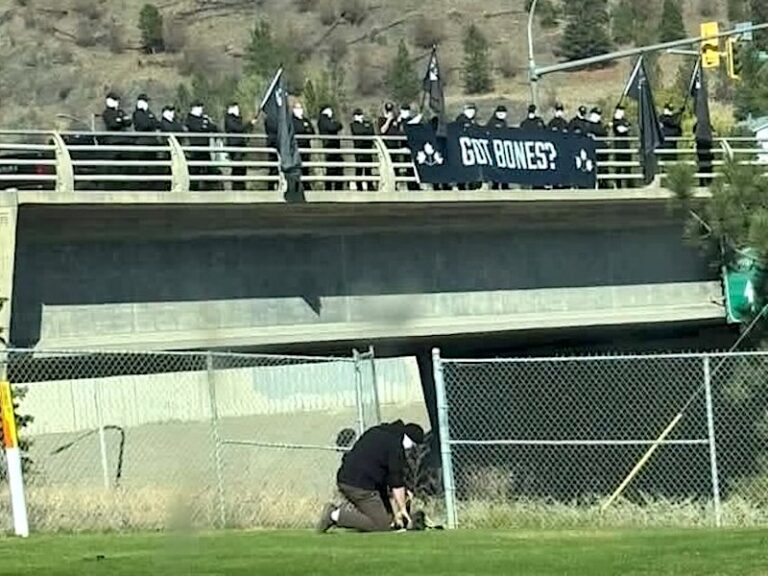 A group of men in black, wearing white masks, stands on a highway overpass holding flags and a white banner that says ‘Got Bones?’ Another man kneels and pretends to dig in the grass below the overpass.