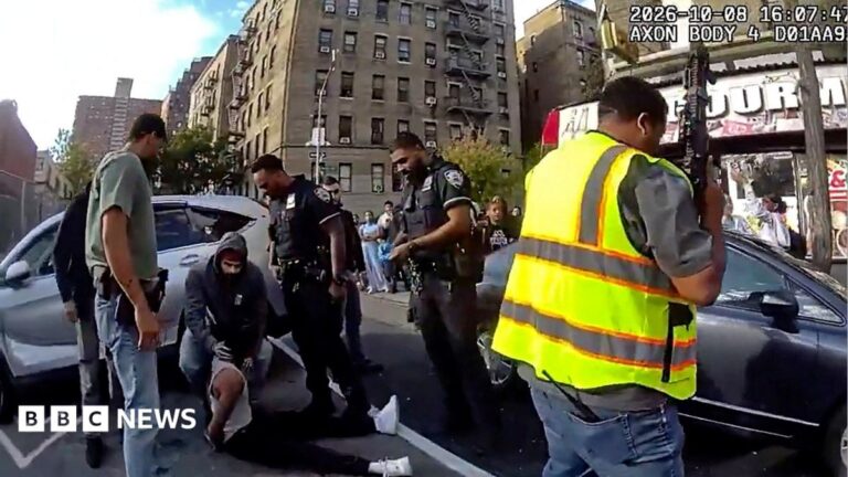 Footage from a New York police officer's body camera shows a man sat on the ground by a silver car with four other men, including police officers, stood around him. One man, wearing a hooded jumper, bends over him and touches his neck. A man holding a large gun and wearing a high-visibility vest is seen in the foreground on the right of the shot, in front of a dark car